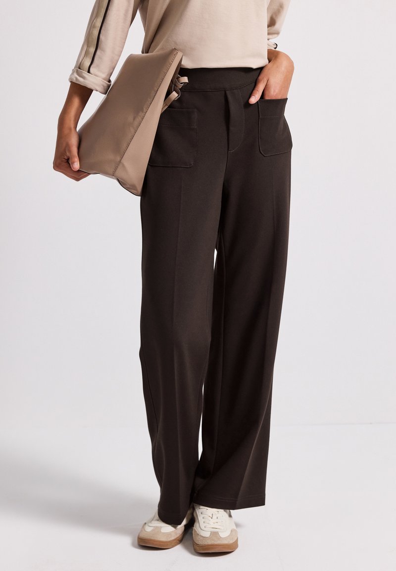 Pantalons larges noirs avec une texture lisse, dotés de deux poches avant. Un sac à main beige est tenu dans la main gauche.