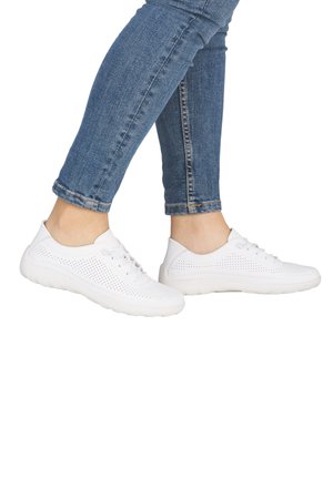 Füße in weißen, perforierten, Schnür-Sneakers und blauen Skinny-Jeans vor weißem Hintergrund.