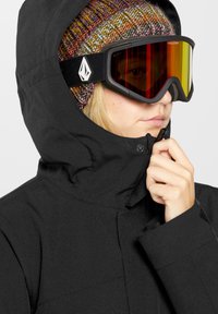 Zwarte winterjas met een hoge kraag, gecombineerd met een meerkleurige gebreide muts en getinte goggles met een logo op de band.