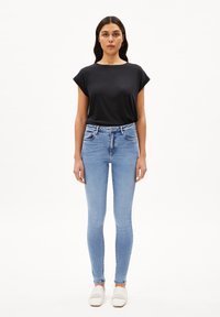 Top negro de manga corta, jeans ajustados de mezclilla azul claro y zapatillas blancas de tejido y sin cordones. Tela suave y diseño ceñido. Fondo blanco.