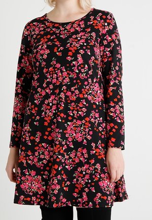 Robe tunique noire à manches longues avec imprimé floral rose vif, rouge et orange portée sur un pantalon noir.