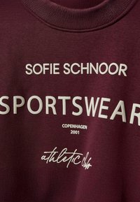 Burgundowa bluza z białym tekstem, który brzmi "SOFIE SCHNOOR SPORTSWEAR COPENHAGEN 2001" oraz "athletic club" w kursywie.