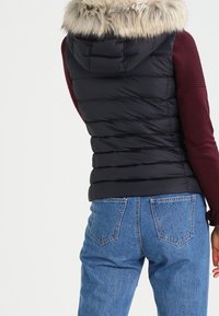 Svart pufferväst med huva i fuskpäls, med stickade ärmar i vinröd färg och horisontell quiltning. Bärs med högmidjade blå jeans.