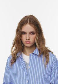 Fată tânără cu păr ondulat de culoare maronie deschisă, purtând o cămașă cu dungi albastre și albe deasupra unor bluze gri și albe, pe un fundal alb simplu.
