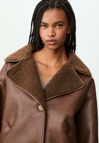 Donna con capelli intricciati, che indossa grandi orecchini a cerchio e un cappotto in pelle foderato di shearling marrone, rivolta di fronte con un'espressione neutra.