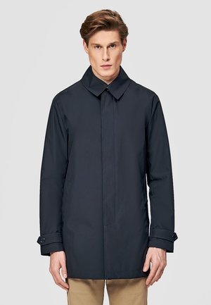 Młody mężczyzna w dopasowanym czarnym płaszczu z zapinanymi na guziki mankietami i beżowych spodniach, stojący na tle jednolitego jasnoszarego tła.