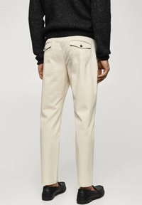 Beige maßgeschneiderte Hose mit glatter Textur, zwei Gesäßtaschen mit Knopfverschluss, getragen mit schwarzen Mokassins. Geeignet für legere oder halbformelle Anlässe.