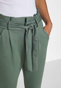 Pantalons légers et verts avec une taille ceinturée et un avant plissé. Possède des poches latérales et une texture douce et lisse.