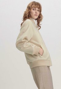 Beige Sweatshirt mit rundem Kragen, Fronttaschen und gerippten Bündchen. Das Material wirkt weich mit einer subtilen Textur. Kombiniert mit hellen Hosen.