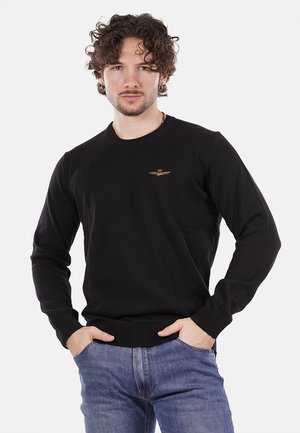 Maglione nero a maglia con scollatura tonda e polsini a costine, caratterizzato da un piccolo emblema dorato sul petto. Abbinato a jeans blu.