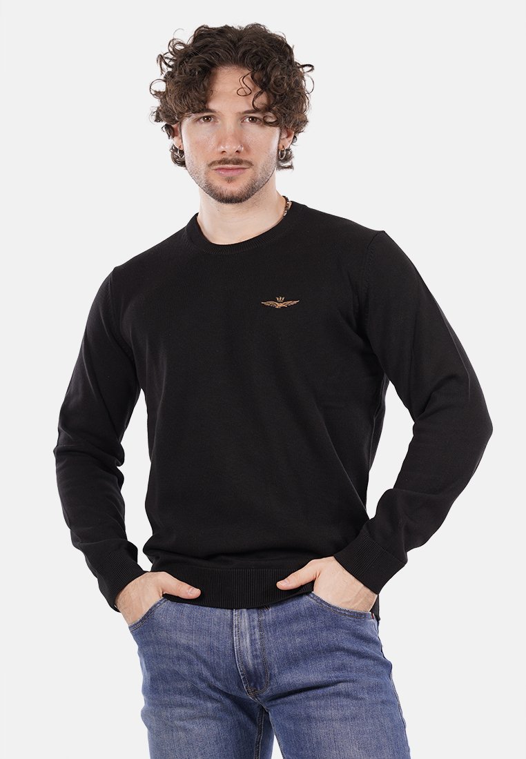Maglione nero a maglia con scollatura tonda e polsini a costine, caratterizzato da un piccolo emblema dorato sul petto. Abbinato a jeans blu.