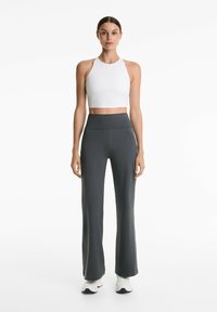 Das Model steht frontal und trägt ein weißes Crop-Top und dunkelgraue Schlaghosen, dazu weiße Sneakers, vor einem schlichten weißen Hintergrund.
