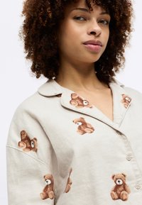Lys beige skjorte med teddy-bjørn broderi. Har knappet front og krage. Myk tekstur med lekne bjørndesign i brune toner.