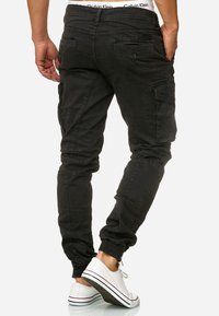Pantaloni cargo neri con una vestibilità affusolata, polsini elasticizzati, molteplici tasche e un tessuto testurizzato. Indossati con sneakers bianche caratterizzate da strisce.
