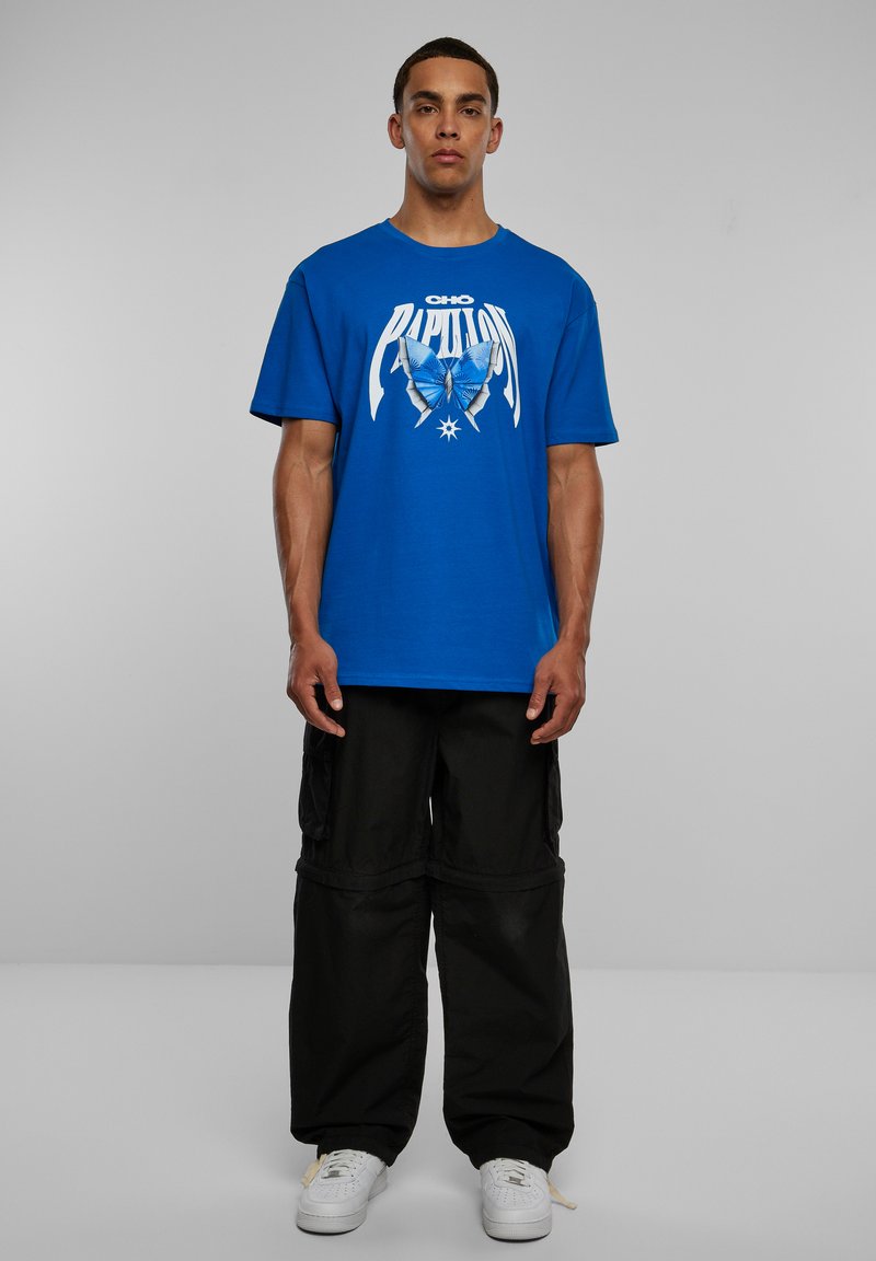 Camiseta de algodón azul con gráfico de mariposa y texto blanco. Combinada con pantalones cargo negros y zapatillas blancas. Diseño sencillo, corte relajado.