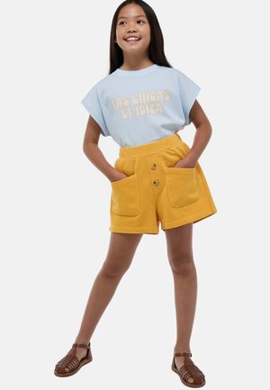 T-shirt bleu clair avec "LAS CHICAS DE IBIZA" en crème, associé à un short jaune doté de deux poches avant et d'une fermeture à trois boutons.