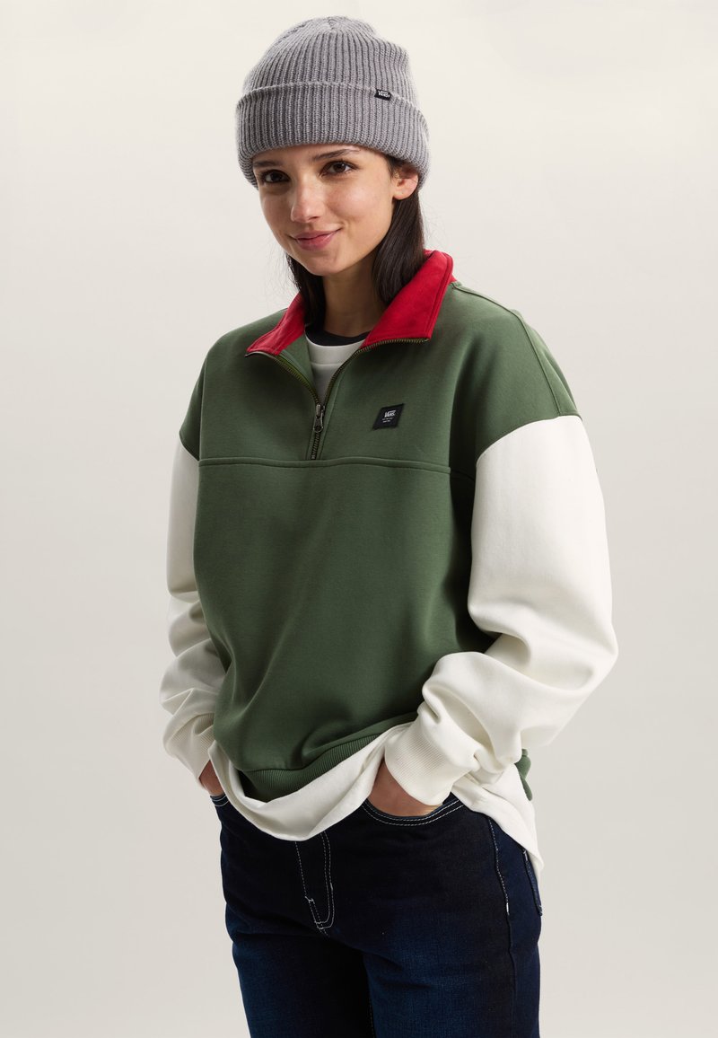 Zelenobílá mikina na zip do poloviny s červeným límcem a černou nášivkou. Kombinováno s tmavými džínami a šedou pletenou čepicí.