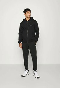 Moški v črni jolki na zadrgo in ustreznih trenirkah s belimi športnimi čevlji, stoji ob enostavni svetli ozadju.