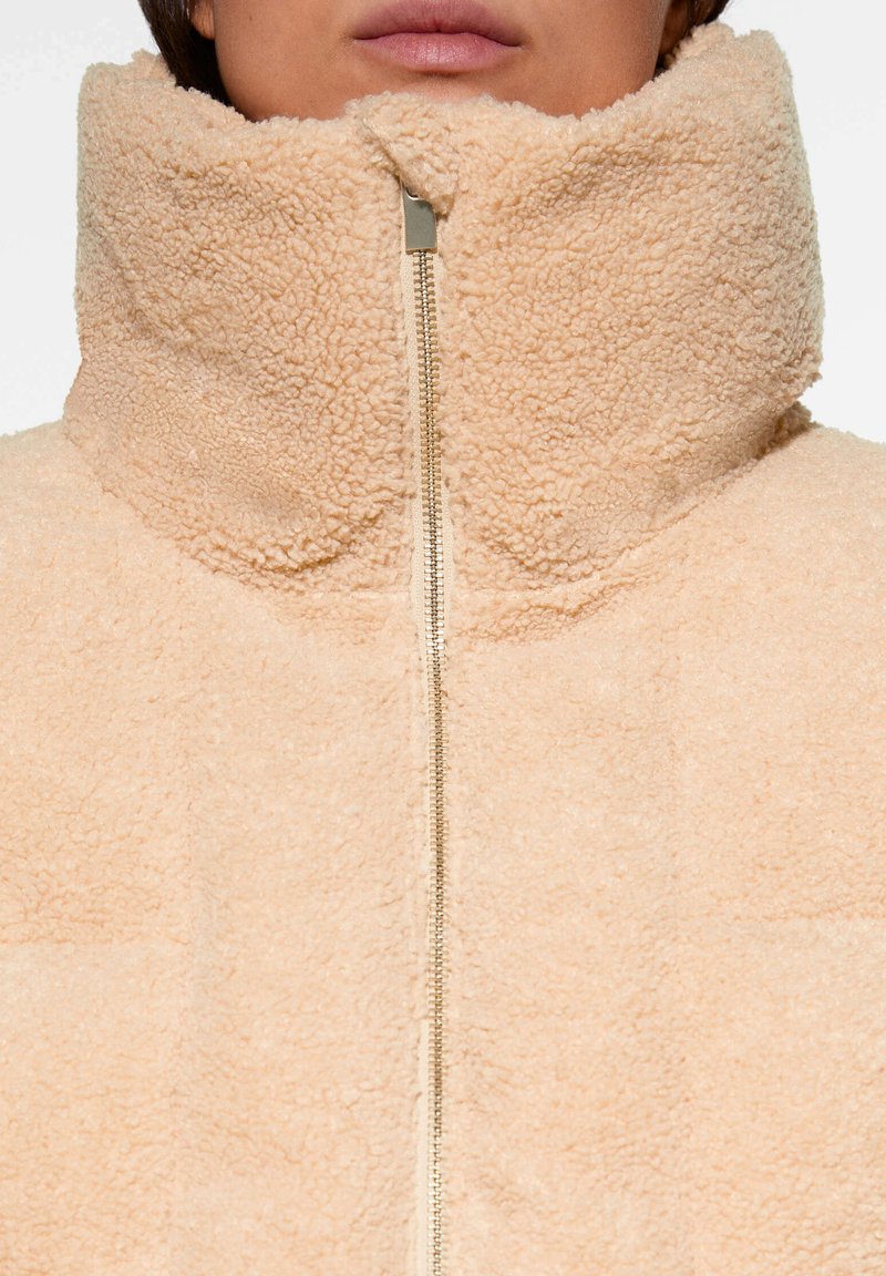 Beige Fleecejacke mit hohem Kragen und einem Front-Reißverschluss. Weiche Textur mit einem leicht flauschigen Aussehen und minimalen Nähdetails.