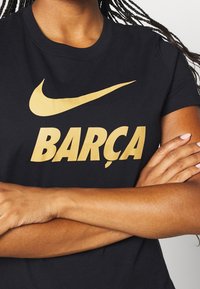 Osoba s copy má na sobě černé tričko s zlatým logem Nike a slovem "BARÇA," paže má zkřížené na hrudi.