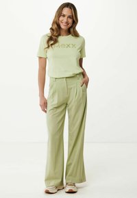 Svetle zelené tričko s grafikou „MXXX“, spárované s vysokými, volnými svetle zelenými kalhotami. Model má na sobě neutrální tenisky.