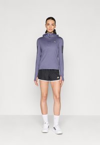 Haut de sport à manches longues violet avec zip moitié et capuche, associé à un short noir avec des accents blancs. Des chaussettes et des chaussures blanches complètent la tenue.