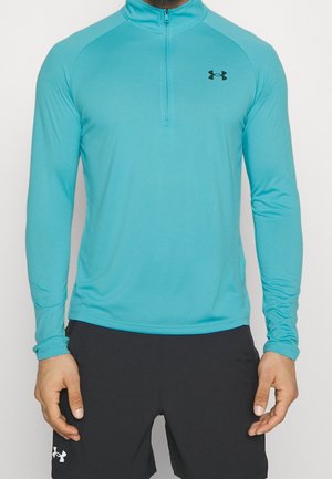 Mężczyzna ubrany w turkusową, długorękawną sportową bluzę Under Armour z półzamkiem i czarne szorty, stojący na jednolitym tle.