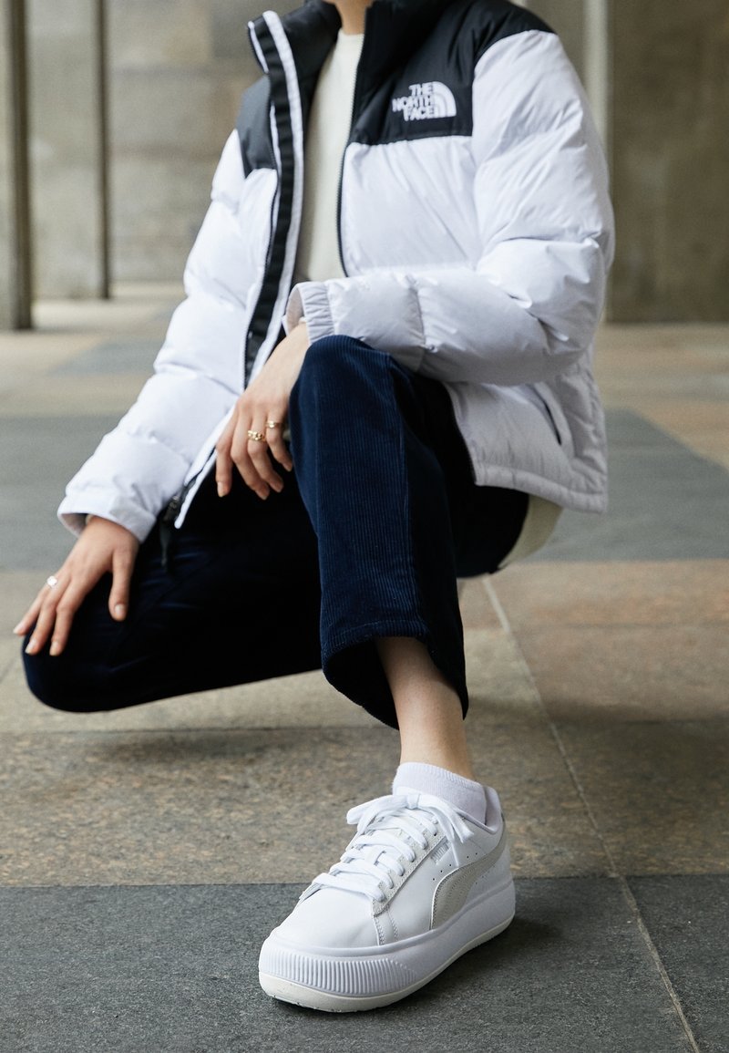 Veste matelassée blanche et noire avec une fermeture éclair, pantalon en velours côtelé bleu marine et baskets blanches avec un logo sur le côté et une semelle texturée, posés sur un sol carrelé.