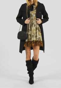 Frau in einem beigen Blumenkleid mit braunem Rüschensaum, schwarzem Mantel, schwarzen lässigen Stiefeln und schwarzer gesteppter Tasche mit goldener Kettenriemen.