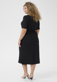 Black ribbed midi dress with short sleeves, side slit, and fitted waist. Worn with nude heeled mules. Curly hair adds volume.