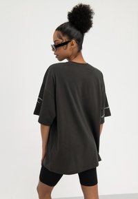 Frau mit lockerer hoher Duttfrisur, die ein übergroßes schwarzes T-Shirt, schwarze Sonnenbrille und schwarze Radlerhosen trägt, posiert vor schlichtem weißem Hintergrund.