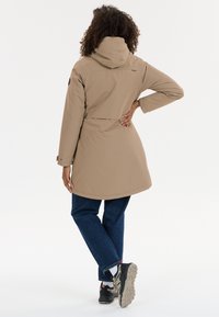 Vrouw met krullend haar draagt een beige gewatteerde jas met capuchon, blauwe spijkerbroek, witte sokken en zwarte sneakers, staand tegen een effen witte achtergrond.