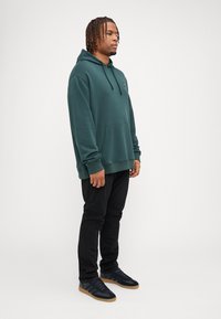 Sudadera de algodón verde con un bolsillo frontal y capucha con cordón, combinada con pantalones negros y zapatillas oscuras con suela de goma.