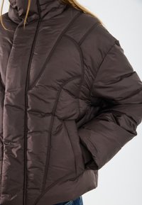 Jachetă pufosă de culoare maro închis, cu cusături texturate, închidere cu fermoar și buzunare laterale, purtată de o persoană cu mâna în buzunar.