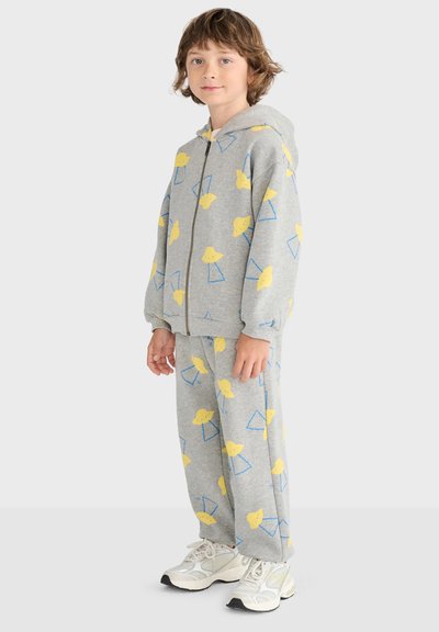 Enfant portant un sweat à capuche gris zippé et un pantalon assorti avec des formes abstraites jaunes et bleues, debout avec des baskets blanches.