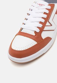 Weiße und braune Leder-Sneaker mit weißen Schnürsenkeln, perforierter Zehenpartie und einem Zickzack-Seitenmuster auf einem weißen Hintergrund.