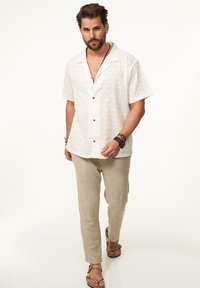 Kurzarmpoloshirt in Weiß mit strukturiertem geometrischen Muster, kombiniert mit hellbeigen gewebten Hosen und braunen Sandalen.