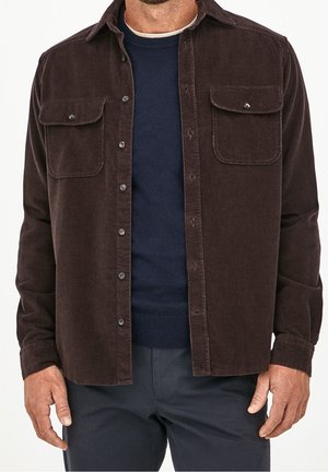 Mężczyzna ubrany w ciemnobrązową sztruksową koszulę zapinaną na guziki, nałożoną na granatowy sweter i ciemne spodnie, stoi z rękami swobodnie opuszczonymi wzdłuż ciała.