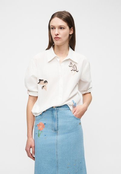 Ung kvinde med glat brunt hår iført en hvid skjorte med svampebroderi og en lyseblå denims nederdel med blomsterbroderi.