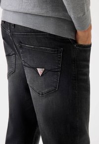 Jeans de denim negre cu croială slim, având o etichetă logo triangulară pe buzunarul din spate și detalii de cusătură vizibile. Textură netedă.