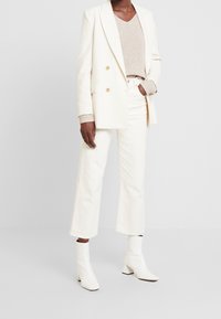 Blazer blanc à boutons croisés porté sur un pull en maille beige, assorti à un jean blanc à jambes larges et des bottines blanches à la cheville.