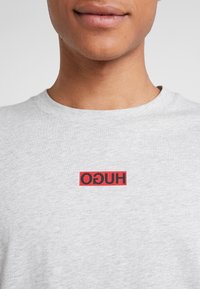 Grijs katoenen t-shirt met een rood rechthoekig logo met "HUGO" in zwarte letters, een ronde hals en korte mouwen.