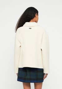 Kooretooniline fliisjope kraega, üle suuruse lõikega ja soonikuga äärega. Kaeluse taga väike brändi silt.