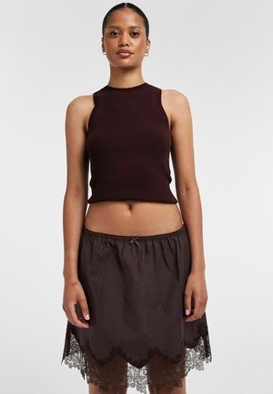 Femme portant un haut court sans manches en maille côtelée de couleur brun foncé et une jupe assortie brun foncé avec des détails en dentelle à l'ourlet, debout devant un fond uni.