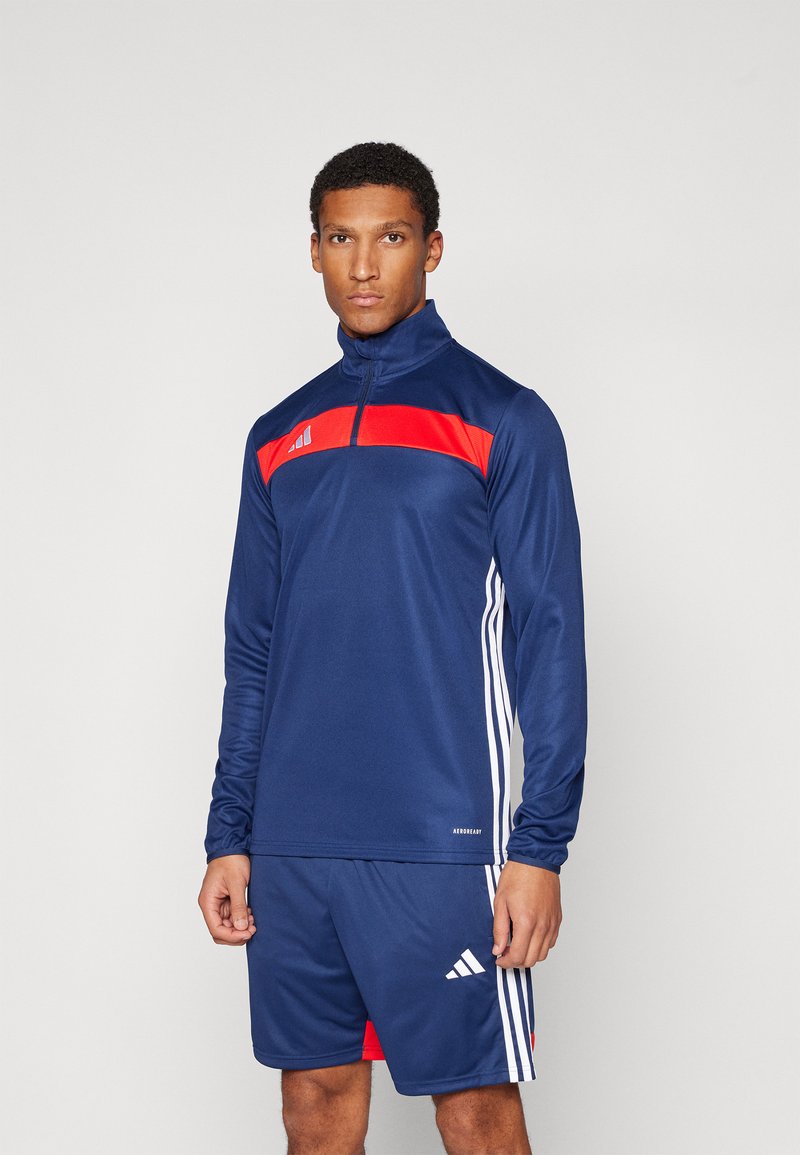 Idrettssett bestående av en marineblå langermet zip-up genser med rød aksentstripe og matchende shorts med hvite sidestreker og logodetaljer.