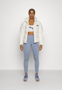 White puffer jacket, crop top with black logo, light blue leggings, and gray sneakers. Model stands against a plain white background.