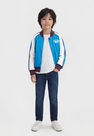Fiatal fiú áll, egyik kezét a zsebében tartva, kék, fehér és piros bomberdzsekit visel, alatta fehér inget, kék farmert és fehér tornacipőt, fehér háttér előtt.