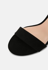 Svart mockasandal med en bred rem över fotryggen och rund tå. Sula i nude-färg som ger kontrast till ovandelen.