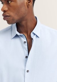 Helles blaues Hemd aus strukturiertem Stoff, mit Knopfkraagen und kontrastierendem marineblauen Besatz entlang der Frontöffnung und des Kragens.