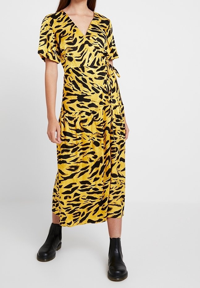 Robe portefeuille jaune à imprimé animalier noir, manches courtes, et ceinture à nouer à la taille. Portée avec des bottines noires.